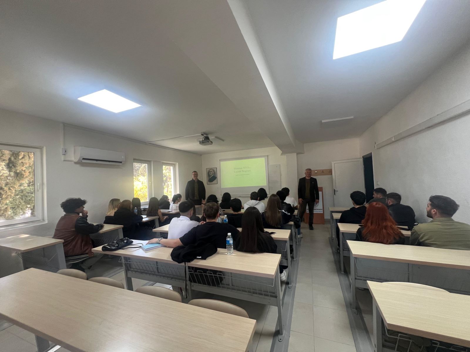 In a classroom setting, students are seated at their desks while two speakers stand in front of the projection screen and give a presentation. The bright classroom includes windows, an air conditioner, and a ceiling-mounted projector.